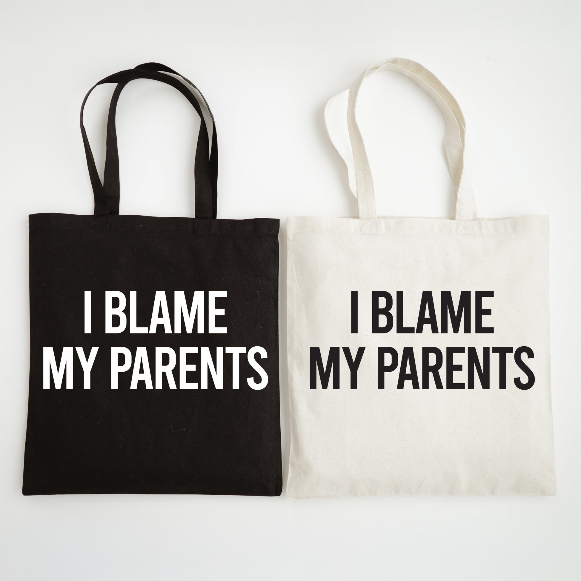 Two tote bags, one black and one white, with 'I Blame My Parents' text on a light gray background.