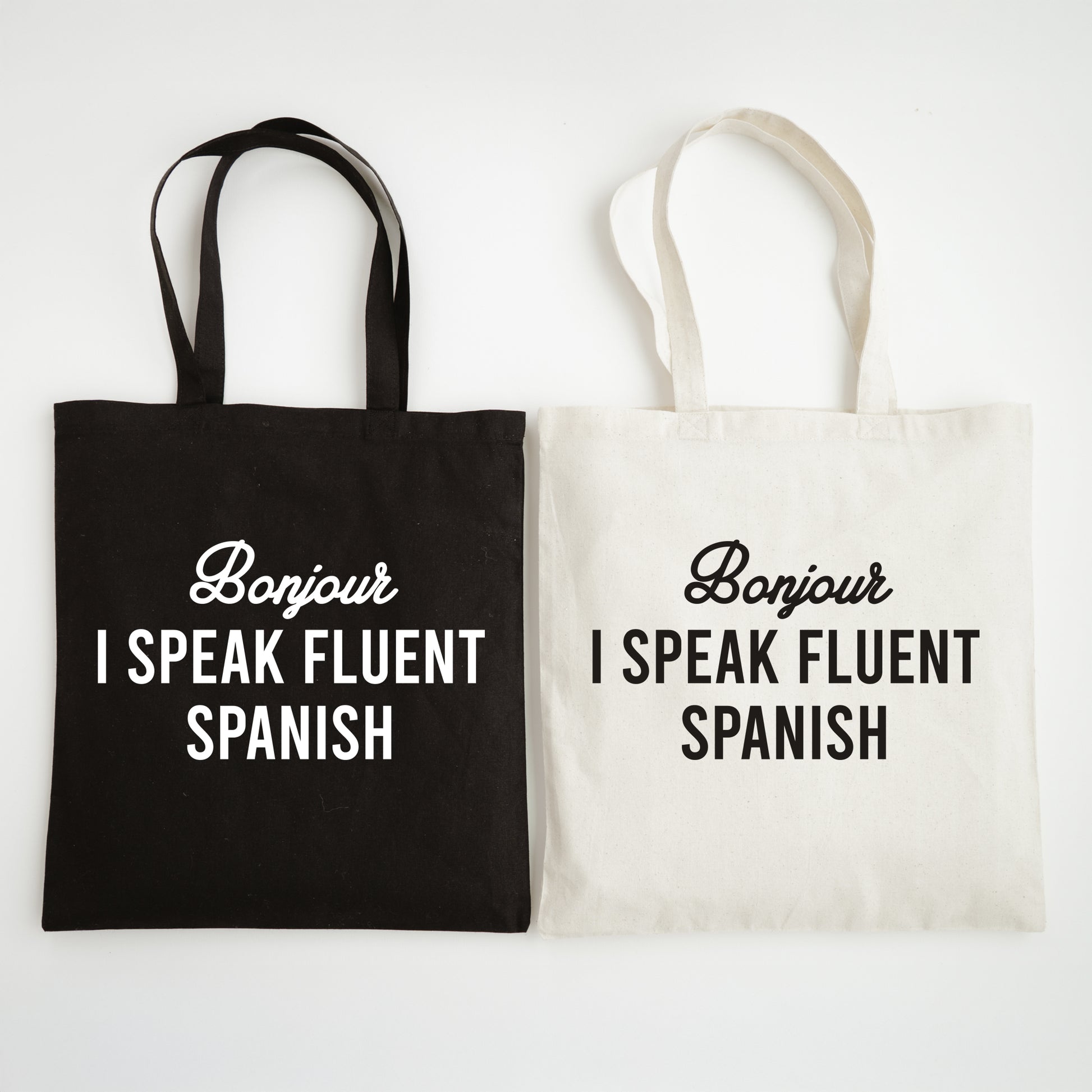 Two tote bags, one black and one white, with text 'Bonjour I speak fluent Spanish' on a light gray background.