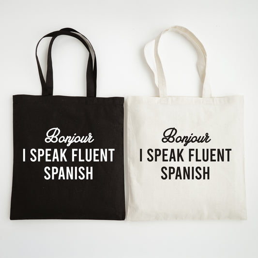 Two tote bags, one black and one white, with text 'Bonjour I speak fluent Spanish' on a light gray background.