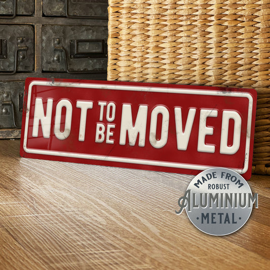 Red sign with 'Not to be moved' text on a wooden surface with wicker basket and metal wall in the background.