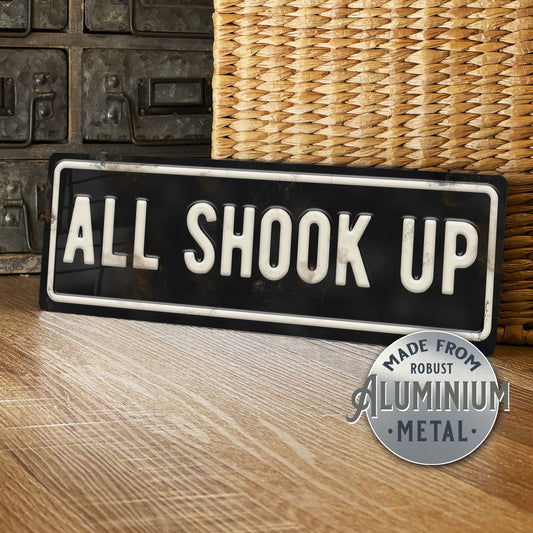 Decorative sign with 'ALL SHOOK UP' text on a wooden surface, with a wicker basket in the background.