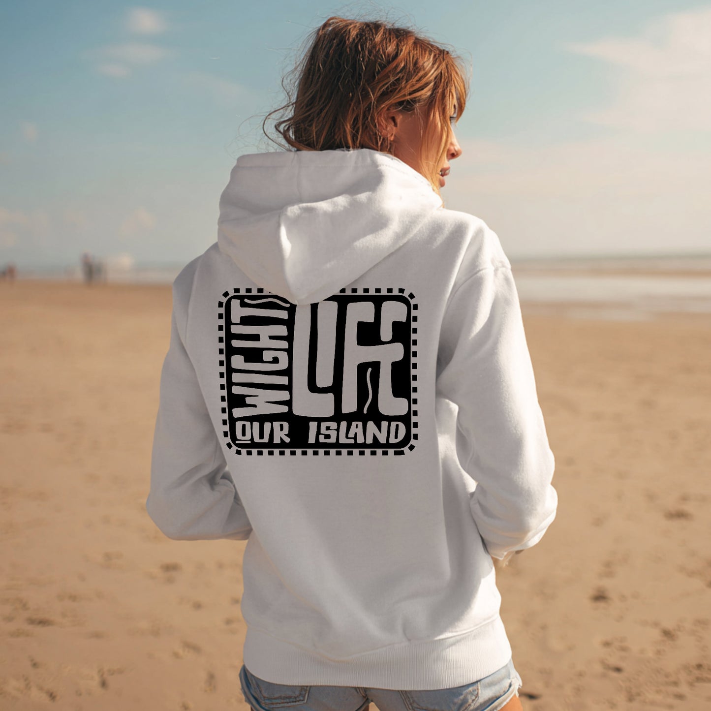 Person wearing a white hoodie with text on the back, standing on a beach.